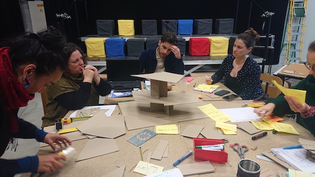 Image de la session de prototypage où l'on peut voir l'équipe en train de construire un jeu de plateau à plusieurs niveaux, à la manière d'un bâtiment de plusieurs étages, avec pleins de post-its et de cartons découpés, au milieu de l'UrbanLab.